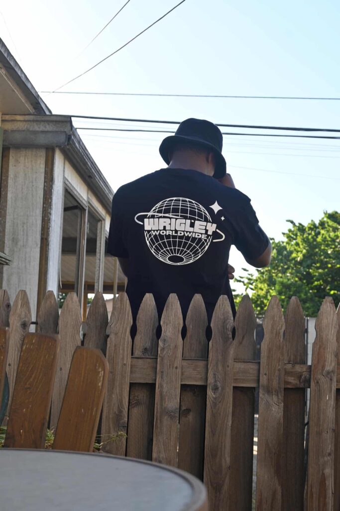 Person in a black hoodie with a graphic logo on the back, standing by a wooden fence.